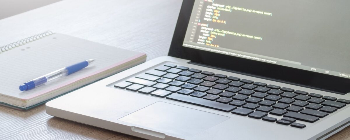 Laptop with code editor open, notebook and pen on wooden desk, perfect for tech and work themes.
