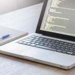 Laptop with code editor open, notebook and pen on wooden desk, perfect for tech and work themes.