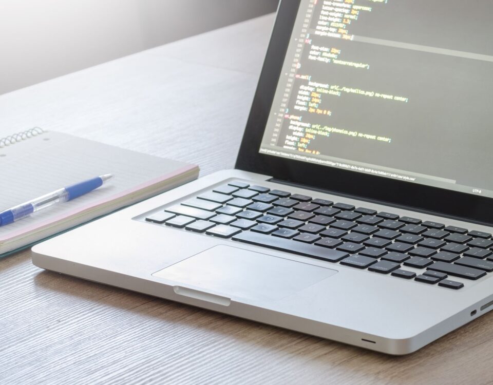 Laptop with code editor open, notebook and pen on wooden desk, perfect for tech and work themes.