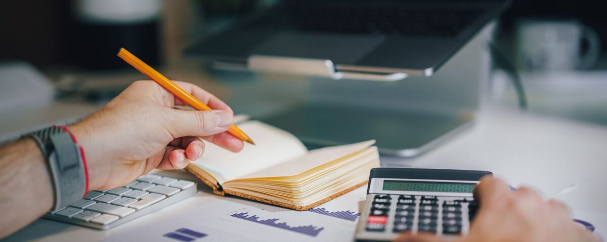 Hands using calculator and taking notes on desk with laptop, showcasing business analytics.