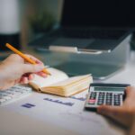 Hands using calculator and taking notes on desk with laptop, showcasing business analytics.