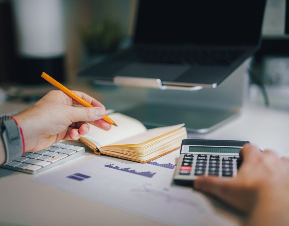 Hands using calculator and taking notes on desk with laptop, showcasing business analytics.