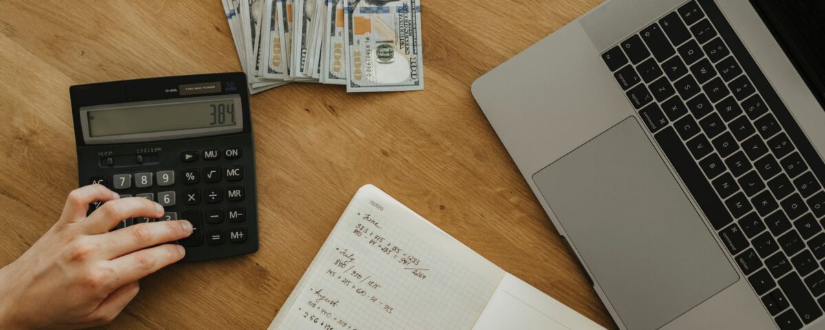 Hands using calculator and notebook, planning finances, with cash and laptop on wooden table.