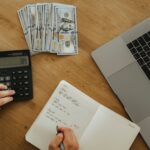 Hands using calculator and notebook, planning finances, with cash and laptop on wooden table.
