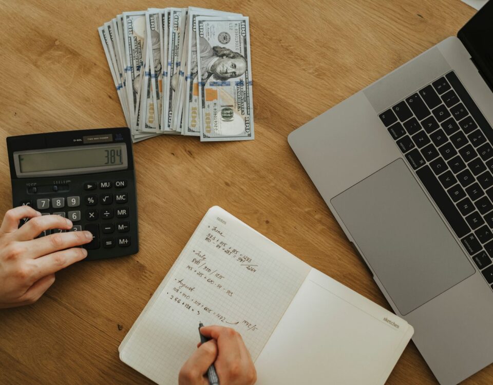 Hands using calculator and notebook, planning finances, with cash and laptop on wooden table.