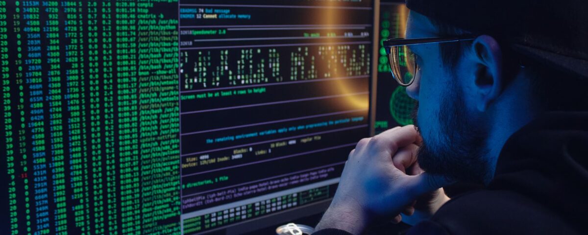 Man intently working on computer programming with code displayed on dual monitors in a dimly lit room.