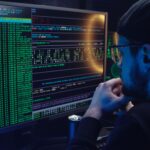 Man intently working on computer programming with code displayed on dual monitors in a dimly lit room.